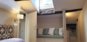 Un dormitorio con cama, lámpara de noche, ventana con vistas y un banco empotrado con almohadones.