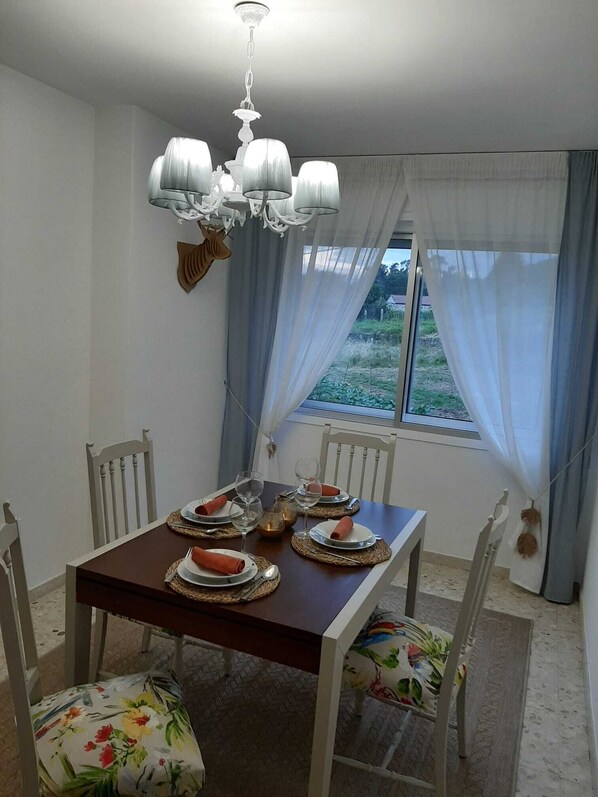 A dining area with a wooden table set for four, white chairs with floral cushions, a chandelier, and a view through a window with sheer curtains. - A CASA DO REGUEIRO, serving wine and thermal water (Cenlle)