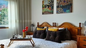 Un lit bien fait, recouvert d’une couette blanche, avec deux oreillers et une table d’appoint sur laquelle se trouvent une tasse et des fleurs. Deux tableaux ornent le mur, il y a une fenêtre avec des rideaux et une table de chevet avec une lampe.