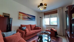 Un salon avec un canapé rouge, une table basse en verre, un tableau au mur, un luminaire au plafond et une fenêtre avec des rideaux.