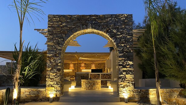 A stone archway leading to a modern outdoor kitchen area with built-in seating and lighting.