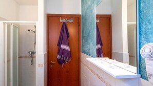Un baño con ducha, una puerta de madera, una toalla colgada en la puerta, un espejo y un lavabo blanco con un perchero para toallas.