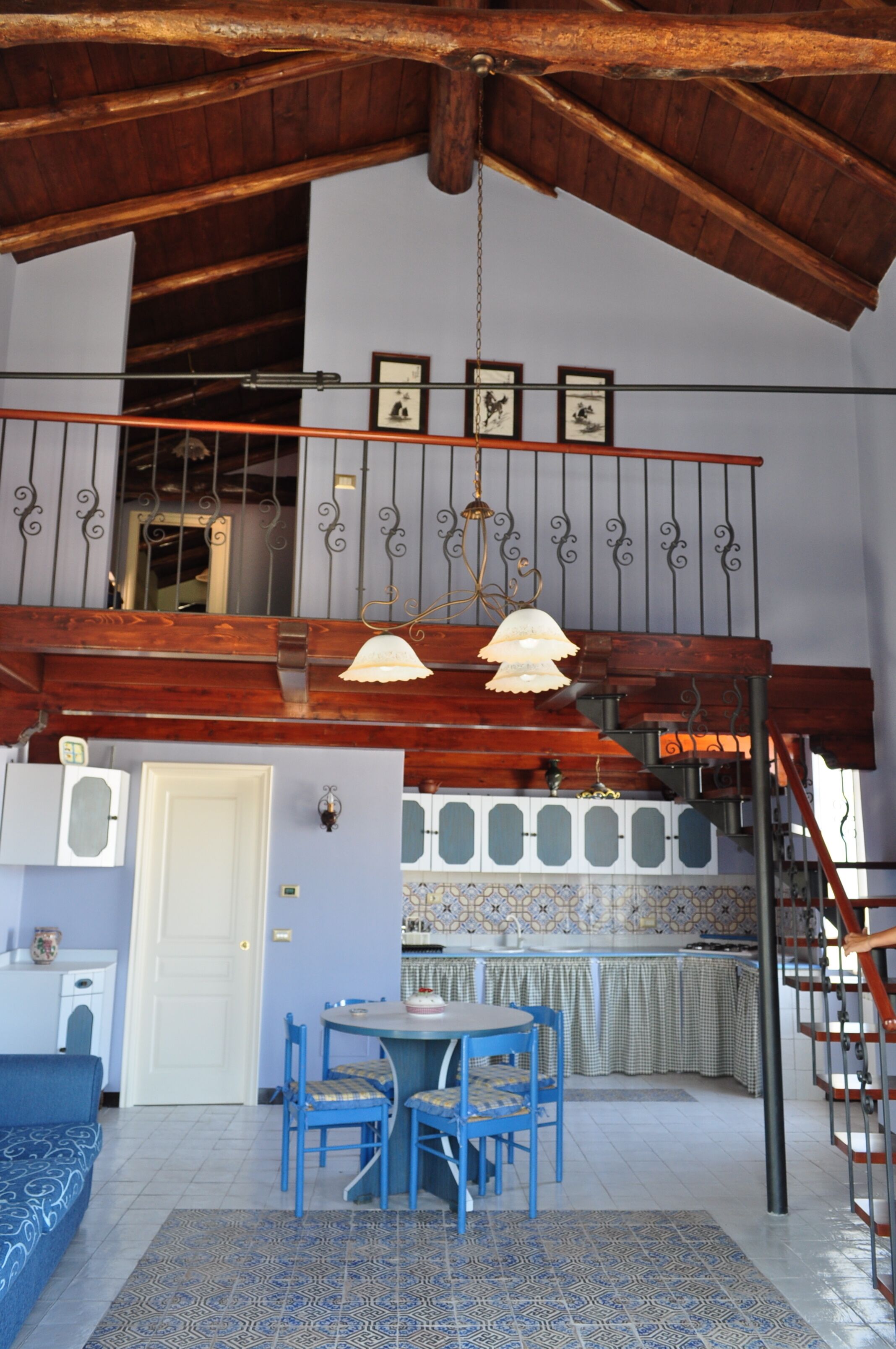 A spacious room with a high ceiling, a dining area with blue chairs, a kitchen with a tiled backsplash, and a staircase with a wrought iron railing.