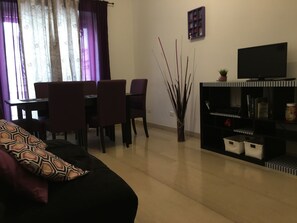 Un salon comprenant un canapé, une table à manger avec des chaises, un téléviseur posé sur un meuble et une plante décorative.