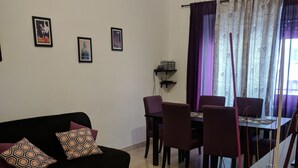 Ein Wohnzimmer mit schwarzem Sofa, violetten Kissen, einem Esstisch mit Stühlen und einem Fenster mit gemusterten Vorhängen.