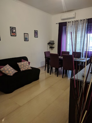 Una sala de estar con un sofá negro, cojines morados, una mesa de comedor con sillas y una ventana con cortinas estampadas.