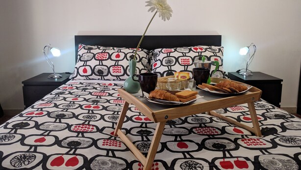 Ein ordentlich bezogenes Bett mit einem gemusterten Bettüberwurf, ein Nachttisch mit einer Lampe und ein Tablett mit Speisen und Getränken auf dem Boden.