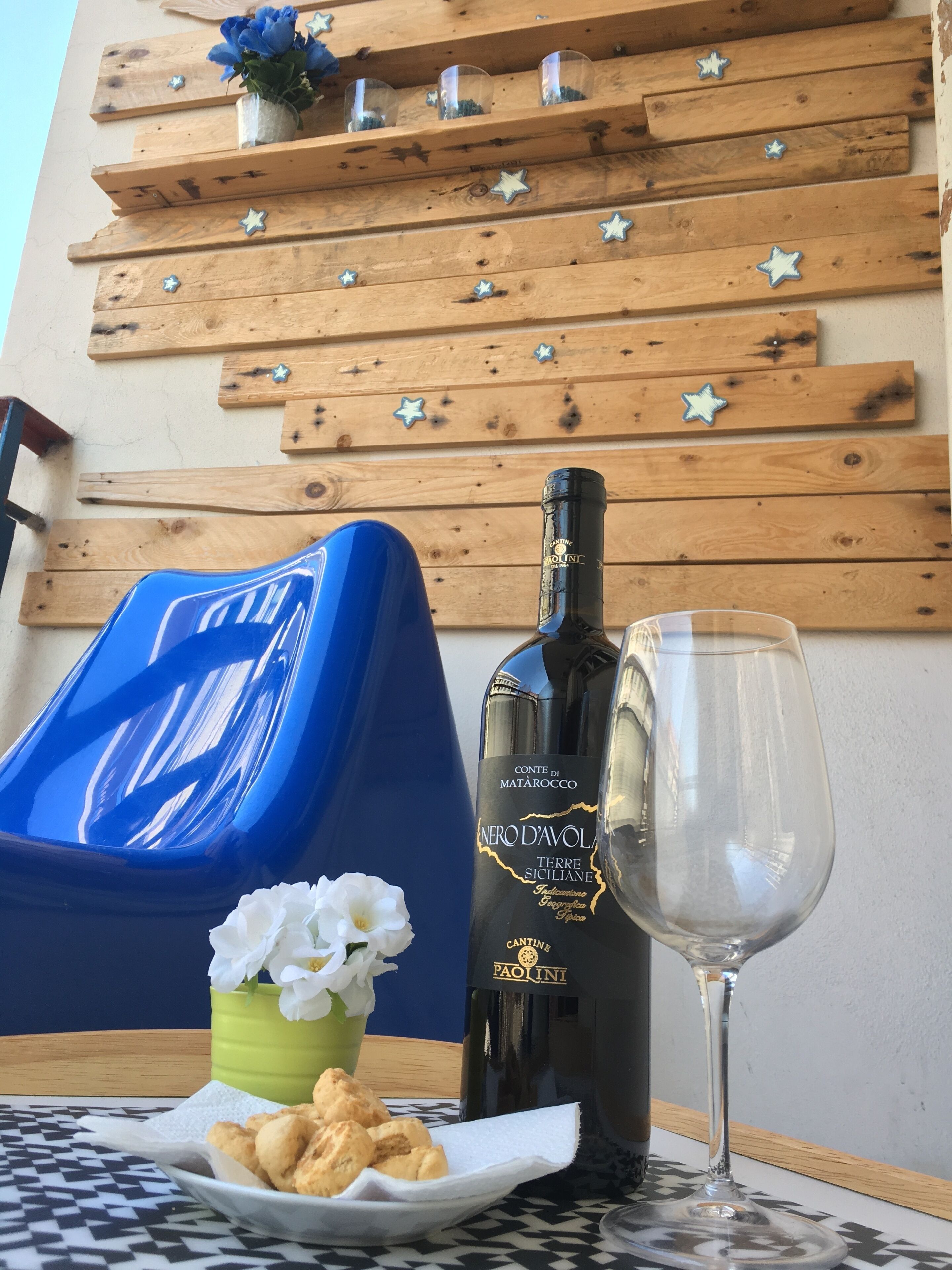 A table with a bottle of NERO D'AVOLA wine, a glass, a small bowl of snacks, and a blue chair.