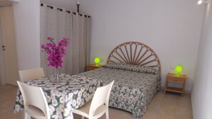 Un dormitorio con cama, mesita de noche con lámpara, una silla, una mesa con mantel floral y un jarrón con flores.