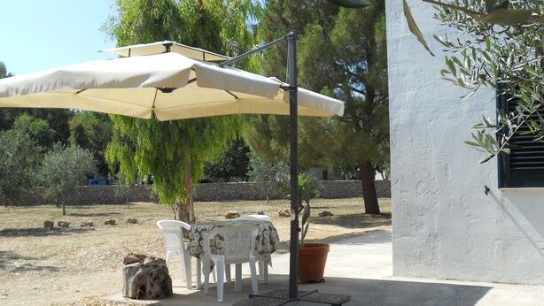 Un espacio al aire libre con mesa y sillas bajo un gran paraguas, rodeado de árboles y un muro de piedra.