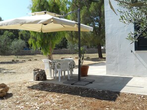 An outdoor seating area with a table and chairs under a large umbrella, surrounded by trees and a stone wall. - Lovely Studio Apartment (Gallipoli (LE))