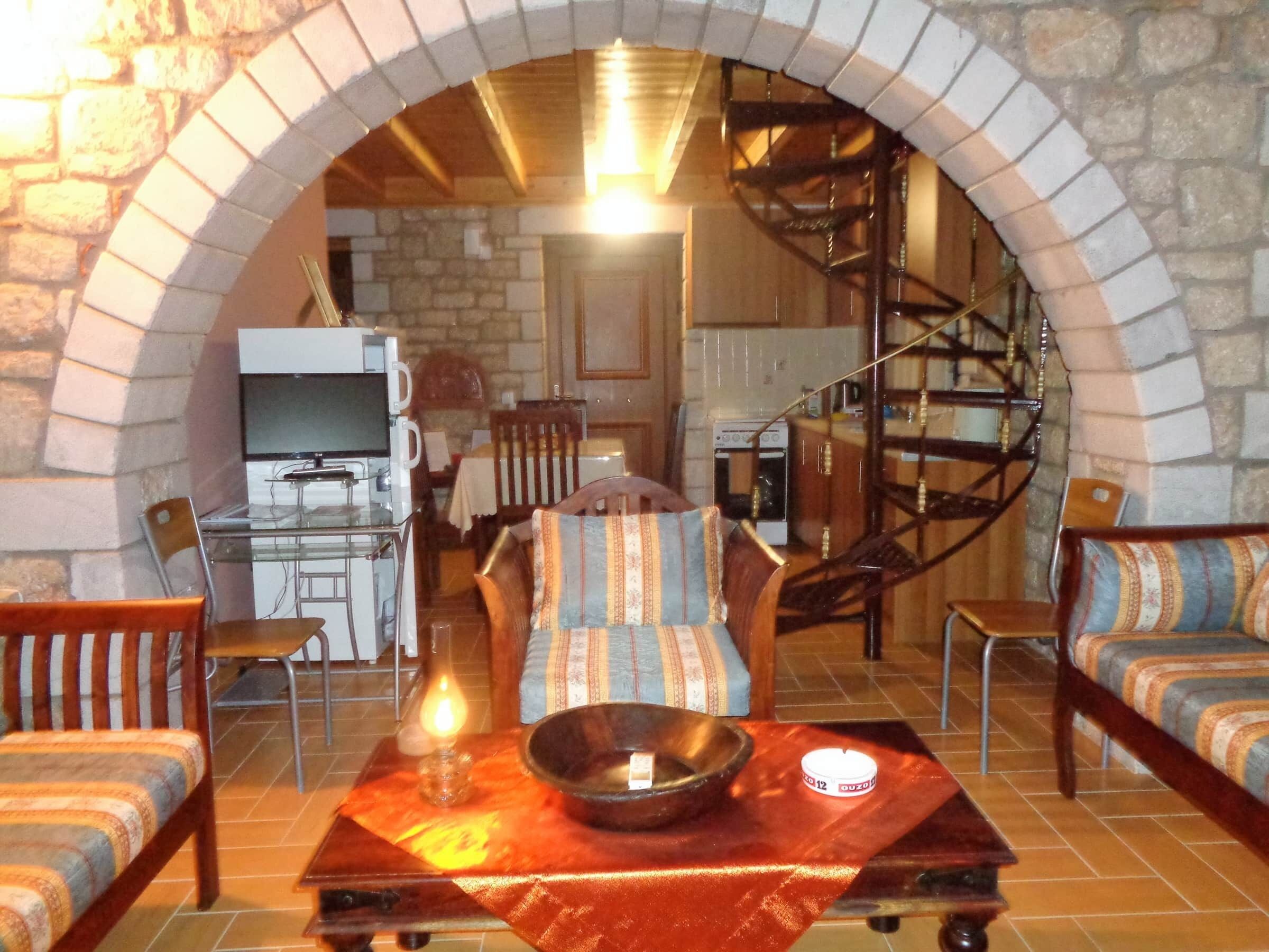 A cozy living room with a stone archway, a dining area with a table and chairs, and a kitchen in the background.