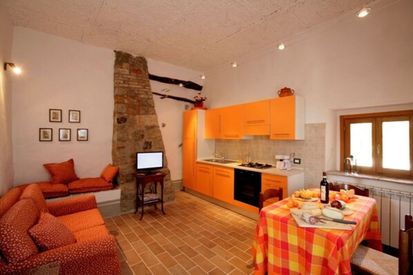 Un espacio habitable compacto con un sofá, una mesa de comedor con mantel a cuadros, una cocina con armarios naranjas y una pared de piedra.