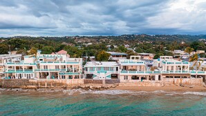 A coastal hotel with multiple buildings, a beach in the foreground, and a hilly landscape in the background.