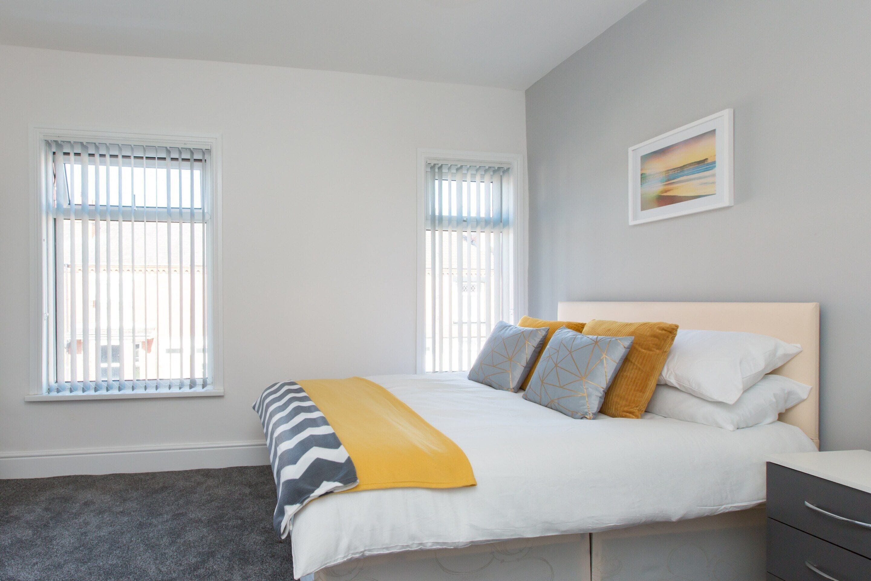 A bedroom with a bed, two windows, a bedside table, and a wall-mounted picture.