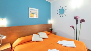 A hotel room with a wooden headboard, an orange bedspread, a framed picture on the wall, a wall clock, and a vase with flowers.