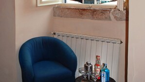 Ett hörnrum med en blå fåtölj, ett litet bord med flaskor och glas, ett element och ett fönster med utsikt över en byggnad med en staty.