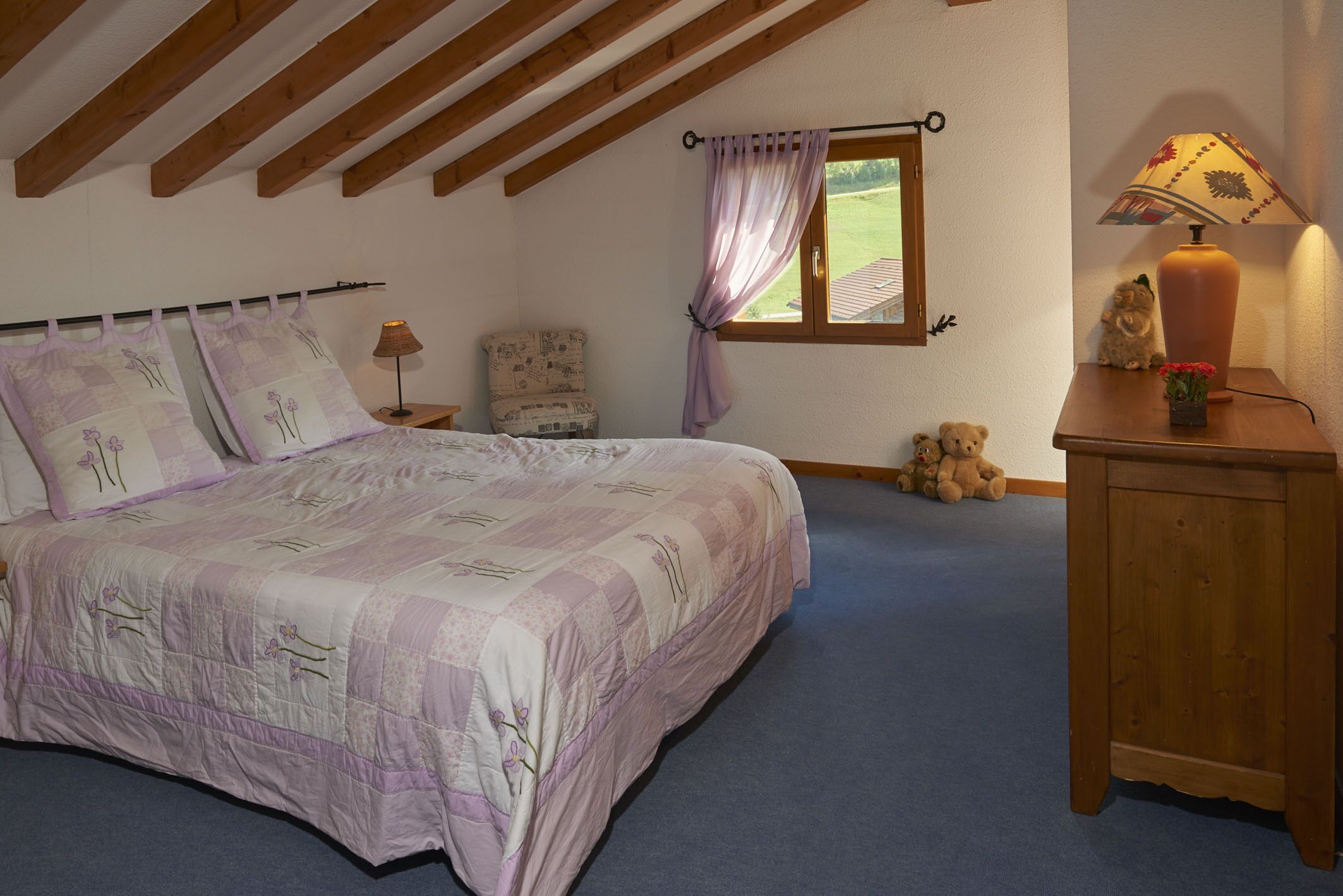 A bedroom with a large bed, a nightstand with a lamp, a chair, a window with curtains, and a teddy bear on the floor.