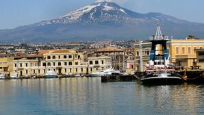 A harbor with boats, a prominent mountain in the background, and a building with a sign reading "CITTÀ DI SIRACUSA."