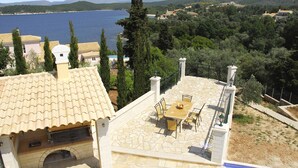 A patio with a stone table and chairs, overlooking a body of water and a mountainous landscape. - Villa Argyro (Corfu)