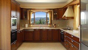 A kitchen with wooden cabinets, a stainless steel refrigerator, and a view of a mountainous landscape through a large window. - Villa Argyro (Corfu)