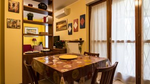 A dining area with a table covered in a floral tablecloth, surrounded by wooden chairs. There are shelves with various items and artwork on the walls.