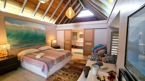 A bedroom with a large bed, a painting of a wave, a desk with a computer, and a view of the bathroom.