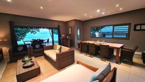 A modern room with a large window overlooking the sea, furnished with wicker sofas, a dining table, and a small kitchen area.