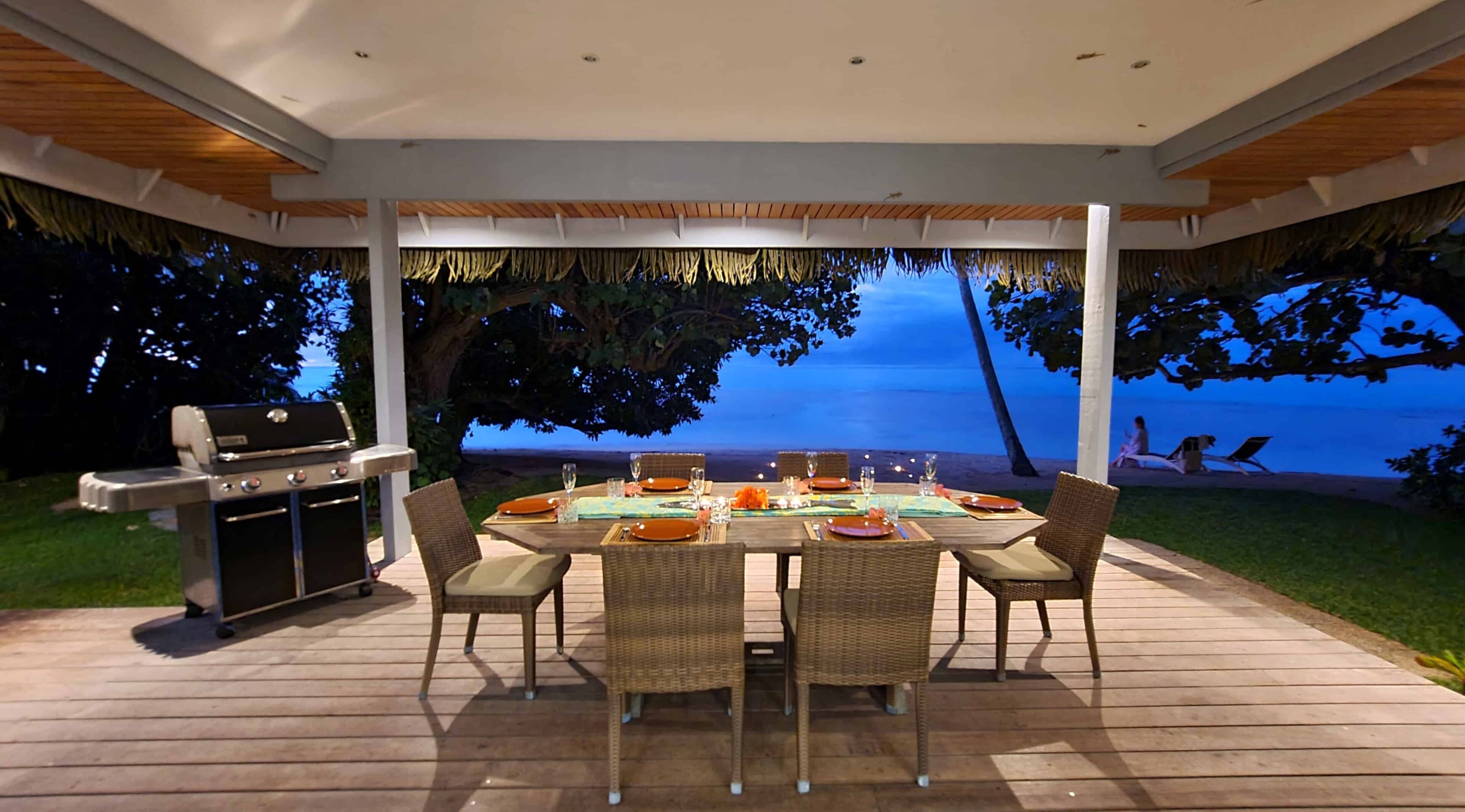 Un espace extérieur aménagé pour les repas, avec vue sur l’océan, un barbecue et des chaises en osier.