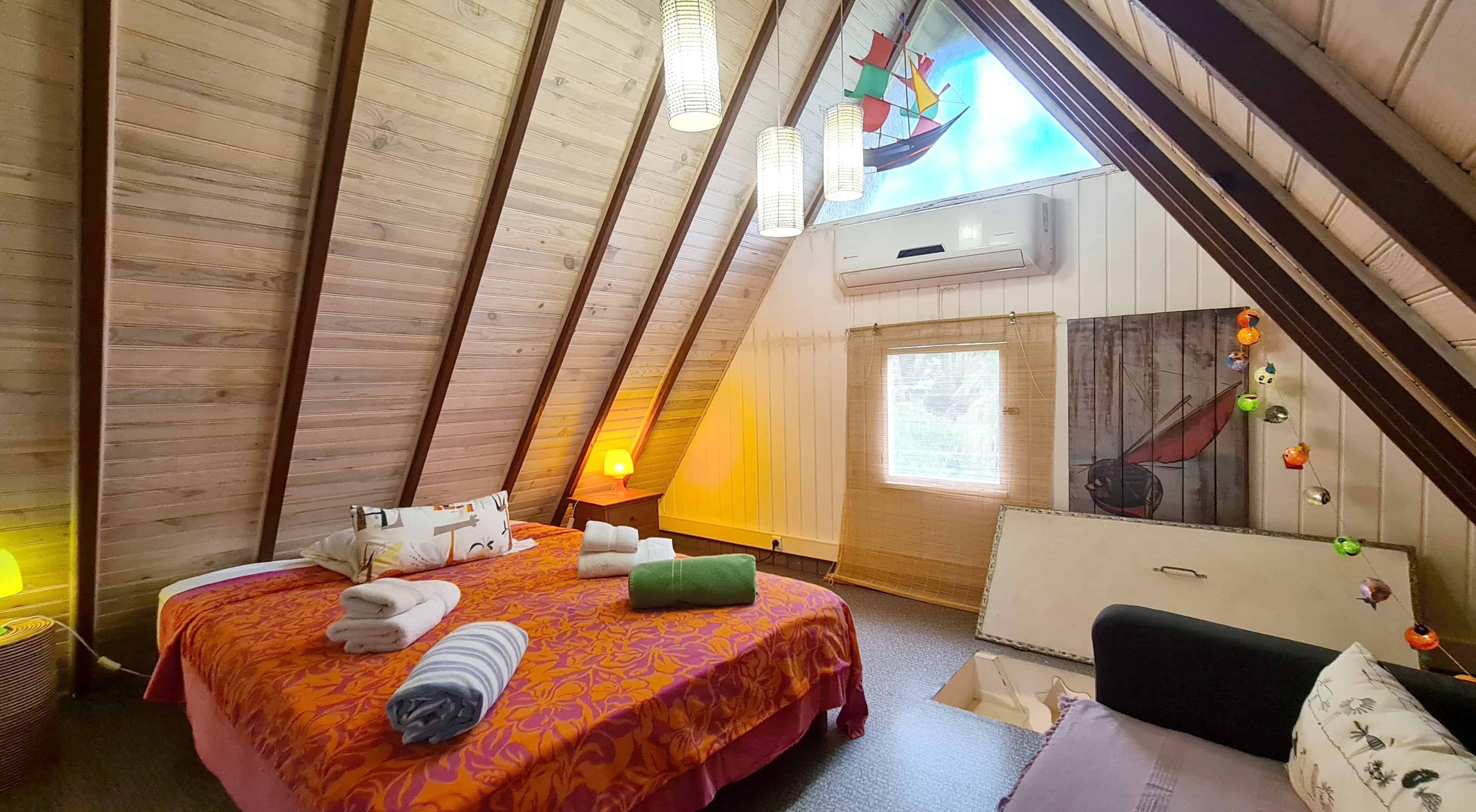 A cozy attic bedroom with a sloped ceiling, a bed with a patterned bedspread, a bedside table with a lamp, and a window with a view of the sky.