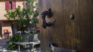 Een houten deur met een metalen handgreep en een metalen plaat, een bank en een boom met witte bloemen.