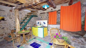 A rustic stone-walled room with a wooden staircase, a small kitchen area, and a dining table with chairs.