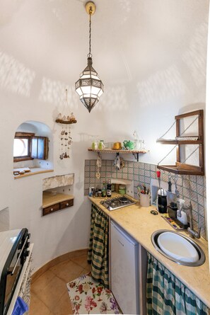 Uma cozinha compacta com luminária pendente, janela com moldura de madeira, pia e pequena geladeira.