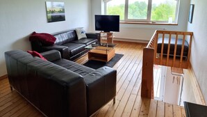 Een moderne woonkamer met een zwarte leren bank, een glazen salontafel, een houten ledikant en een groot raam met uitzicht op groen.