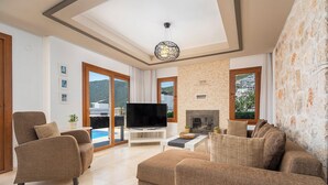 A spacious living room with a flat-screen TV, comfortable seating, a fireplace, and large windows offering a view of the outdoors.