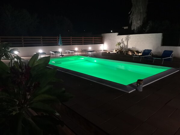 An outdoor swimming pool with a green-lit edge, surrounded by lounge chairs and a wooden deck. - La Rosa Rossa apartment in villa with garden and wonderful panoramic view (Sciacca, Agrigento)