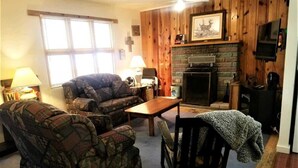 A living room with a fireplace, wooden walls, a sofa, a chair, and a television. - Rustic Cabin (Ruidoso)