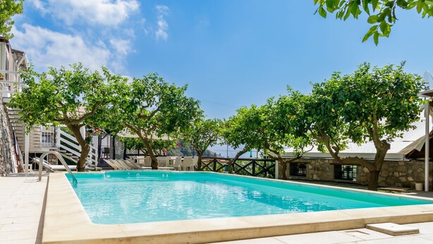 A rectangular swimming pool with a stone deck, surrounded by trees and a stone building.