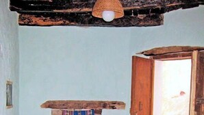 Une pièce avec un sol carrelé, une table en osier recouverte d’une nappe colorée, une chaise en osier, une petite cheminée avec une couverture à rayures et une porte en bois ouverte donnant sur l’extérieur.