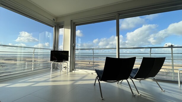 A balcony with a TV, two chairs, and a view of the beach and ocean.