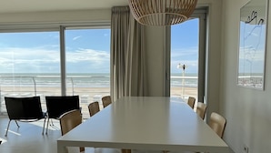 A modern dining room with a large table and chairs, large windows with a view of the beach, and a unique hanging light fixture.