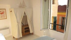 Ein Schlafzimmer mit einem Bett, einem Fenster mit Blick auf Gebäude, einer kleinen Holzablage und einem weißen Baldachin.