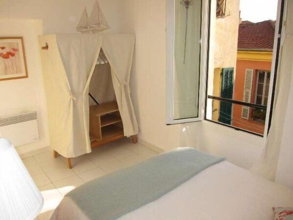 A bedroom with a bed, a window with a view of buildings, a small wooden shelf, and a white canopy.