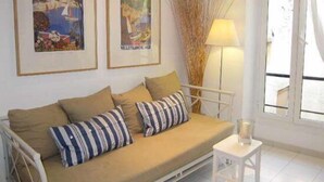 Ein Zimmer mit einem beigefarbenen Sofa, zwei gerahmten Bildern an der Wand, einer hohen Vase mit Ästen, einer weißen Lampe und einem Fenster mit Vorhängen.