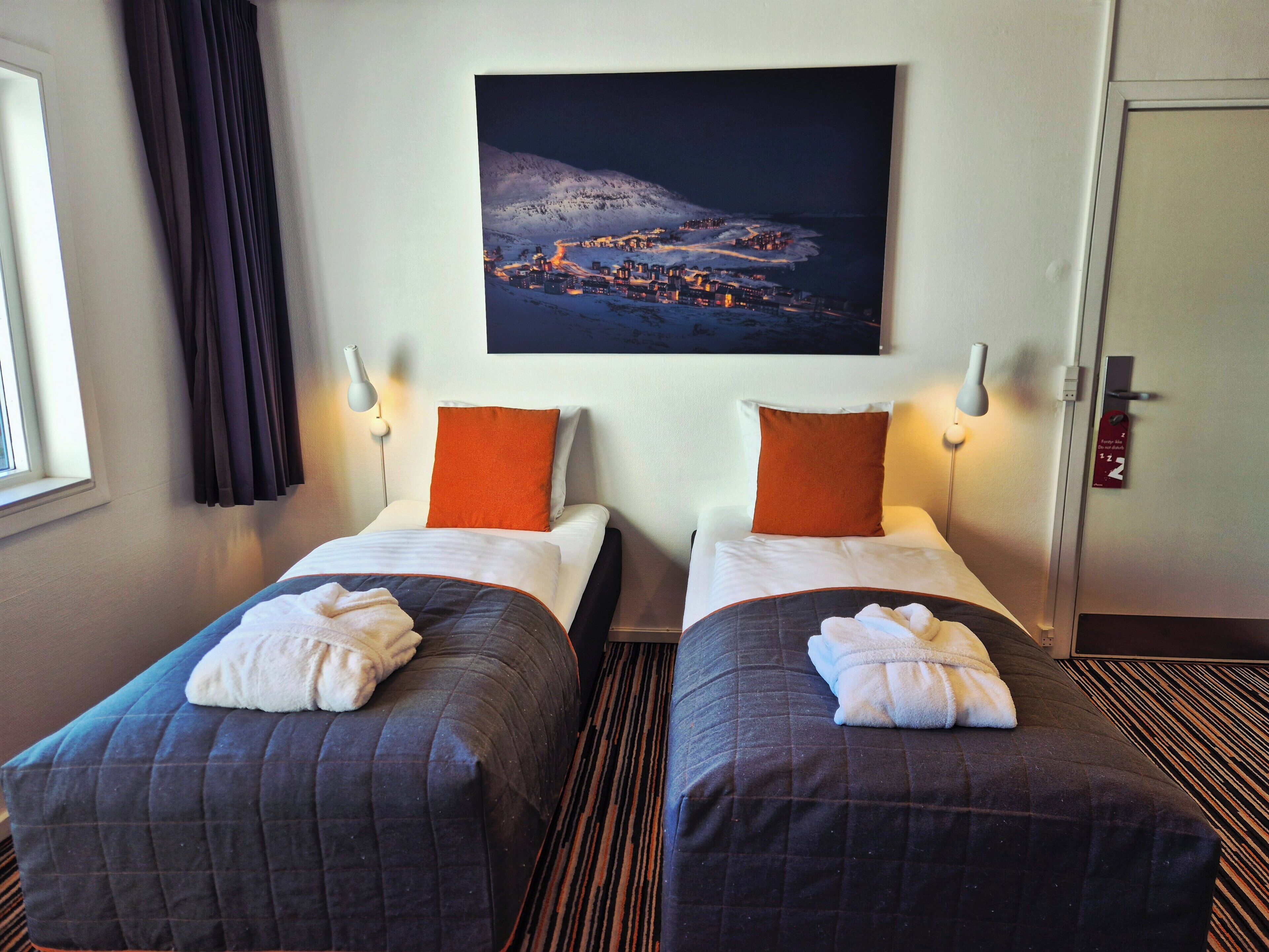 Una habitación de hotel con dos camas, cada una con una almohada naranja y una toalla blanca. En la pared, sobre las camas, hay un cuadro de un paisaje nevado.