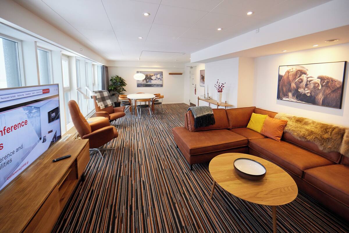 A modern hotel lobby with a large screen displaying conference details, a seating area with a sofa and chairs, and a dining table with a plant.