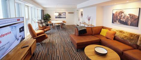 A modern hotel lobby with a large screen displaying conference details, a seating area with a sofa and chairs, and a dining table with a plant.