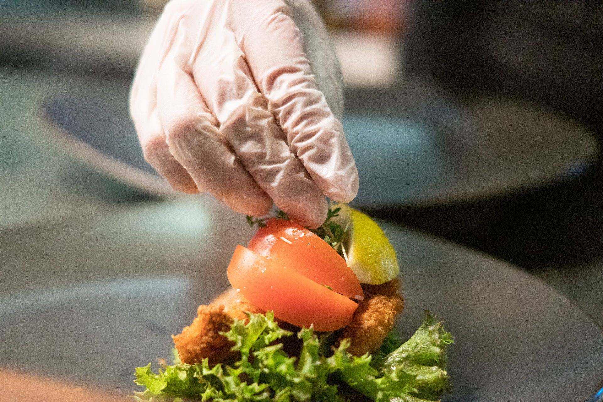 A gloved hand placing a tomato on a dish with lettuce and a lemon slice.