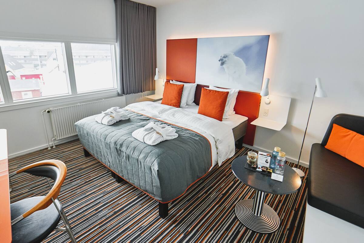 A hotel room with a bed, a chair, a small table with toiletries, and a view of buildings outside.