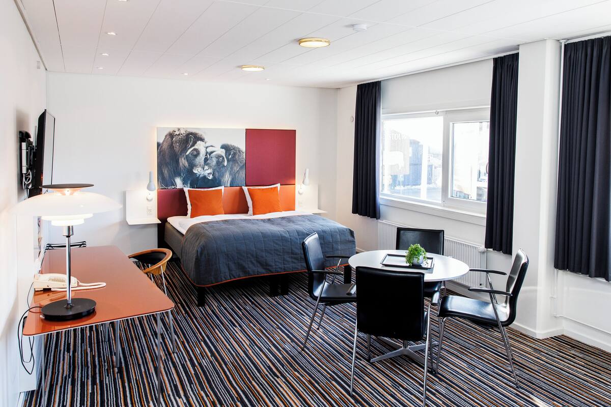 A hotel room with a bed, a desk with a lamp, a chair, a round table with a plant, and a window with curtains.